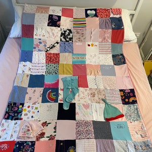 May include: A patchwork quilt made from baby clothes, with squares of different colours and patterns. A pair of light blue tights, a pink floral hat, and a green and red striped hat are placed on the quilt. The quilt is on a bed.