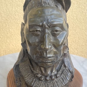 May include: A bronze-toned bust sculpture depicting a person with a topknot hairstyle, wearing a beaded necklace and a fringed collar. The sculpture is mounted on a circular wooden base with a visible grain pattern, showcasing detailed facial features.