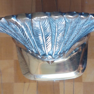 Puede incluir: Un cuenco decorativo plateado con un borde en forma de abanico con detalles en forma de pluma. El cuenco tiene una base rectangular y una superficie pulida y reflectante. El dise&ntilde;o es ornamentado y detallado.