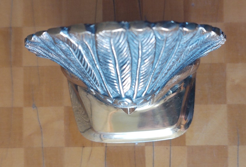 Puede incluir: Un cuenco decorativo plateado con un borde en forma de abanico con detalles en forma de pluma. El cuenco tiene una base rectangular y una superficie pulida y reflectante. El dise&ntilde;o es ornamentado y detallado.