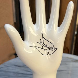 May include: A glossy white ceramic hand-shaped jewelry display stand featuring a black line-art illustration of a bird on the palm. The base of the stand is inscribed with the black text, A Bird in the Hand, in a cursive script font.