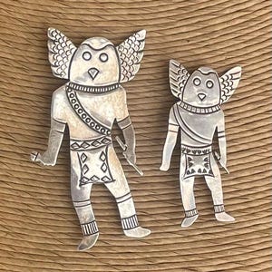 May include: Two silver-colored, stylized figures with owl-like features and wing-shaped elements. The figures have detailed designs on their clothing and are set against a textured, brown background. One figure is larger than the other.