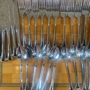May include: A collection of polished silverware, including knives, forks, and spoons, arranged on a wooden surface. The silverware has a reflective, metallic appearance, with detailed handle designs. The arrangement suggests a formal dining setting.