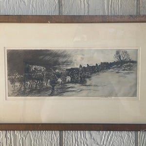 May include: A framed black and white print depicting a line of covered wagons and figures crossing a body of water. The artwork is in a rectangular wooden frame, with a light-colored matting. The print has a vintage aesthetic.