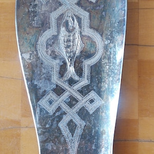 May include: Silver-toned serving utensil with an ornate fish and geometric design. The vintage piece has a decorative handle and a wide, slightly curved blade. The design is etched into the metal, adding texture and detail to the serving piece.