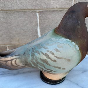May include: A decorative pigeon figurine featuring a dark brown head and neck, transitioning into a light blue and grey body with mottled patterns. The bird has a small white beak, a dark eye, and is mounted on a circular black base.