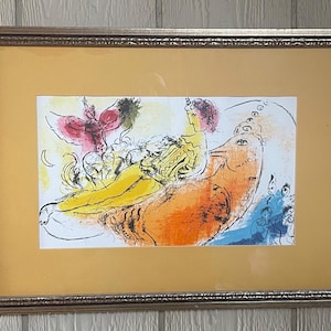 May include: A framed print featuring a colorful abstract illustration. The artwork depicts a figure in yellow and orange, with a red winged figure above. The print is set against a yellow background and framed in a gold-toned frame.
