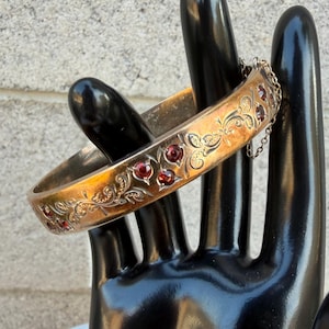 May include: A gold-toned hinged bangle bracelet featuring an engraved floral scroll pattern and inlaid deep red circular gemstones. The bracelet includes a thin metal safety chain attached to the side, providing a secure closure mechanism.