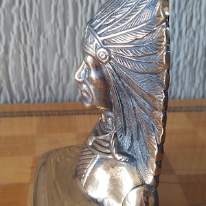 Puede incluir: Escultura de color lat&oacute;n de un jefe nativo americano de perfil, con un tocado de plumas detallado. La escultura tiene un acabado pulido y est&aacute; montada sobre una base rectangular. Los detalles de la cara y las plumas son claramente visibles.