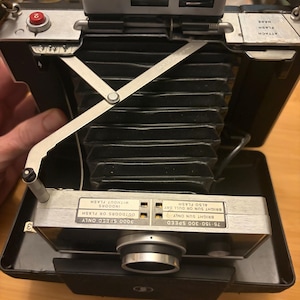 May include: A vintage folding camera with a black accordion bellows, a silver top housing, and a silver lens assembly. The camera features printed exposure instructions, a red shutter button, and a metal support arm. Text includes ATTACH FLASH HERE.