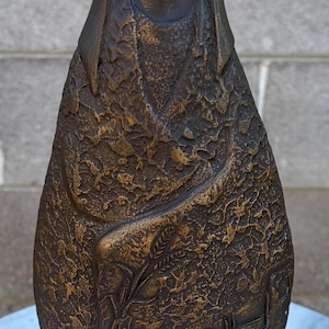 May include: A bronze-toned sculpture featuring a stylized figure with an upward-gazing face. The body is draped in a textured garment adorned with relief carvings of wheat stalks and a small building structure, finished in a dark metallic patina.