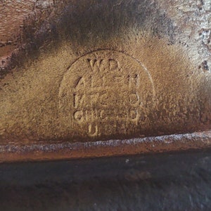 Puede incluir: Primer plano de una superficie de cuero marr&oacute;n desgastado con un logotipo circular en relieve. El logotipo dice "W.D. ALLEN MFG. CO. CHICAGO U.S.A." en un estilo vintage, lo que sugiere un art&iacute;culo antiguo.