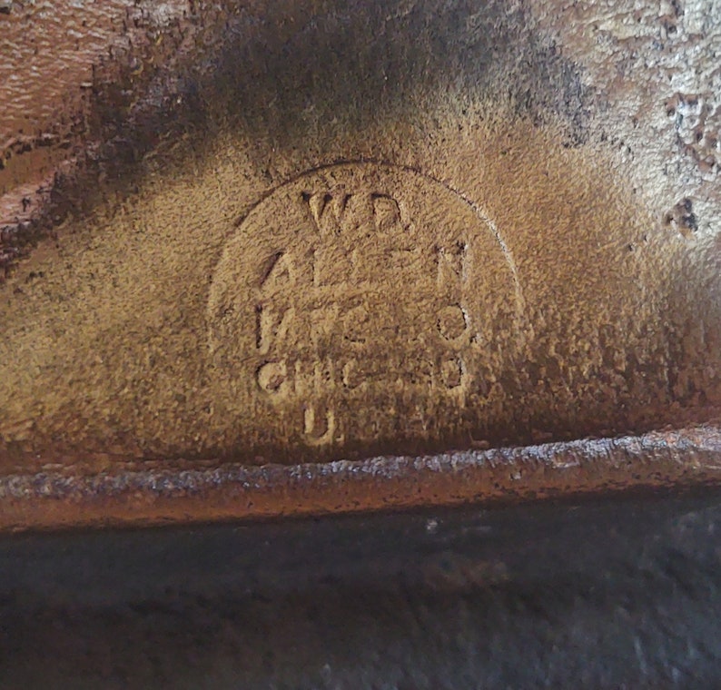 Puede incluir: Primer plano de una superficie de cuero marr&oacute;n desgastado con un logotipo circular en relieve. El logotipo dice "W.D. ALLEN MFG. CO. CHICAGO U.S.A." en un estilo vintage, lo que sugiere un art&iacute;culo antiguo.
