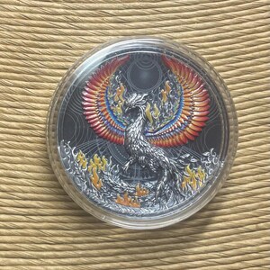 May include: A silver coin featuring a detailed phoenix design with vibrant red, orange, and blue wings. The coin is encased in a clear protective case, set against a textured background.