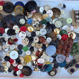 May include: A large collection of assorted sewing buttons in various shapes, sizes, and colors, including metallic, plastic, and wooden materials. Some buttons are loose on a white tray, while others are attached to cardboard display cards.