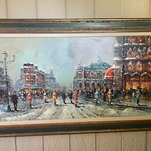 May include: Oil painting of a snowy European city scene, framed in a green and gold wooden frame. The artwork depicts a street with buildings, people, and a lamppost. The buildings have lights on, and the sky is overcast.