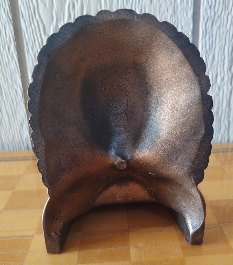Puede incluir: Cenicero decorativo de color bronce con borde festoneado. El cenicero tiene forma de cuenco poco profundo y est&aacute; sostenido por tres patas. La superficie tiene un aspecto ligeramente texturizado.