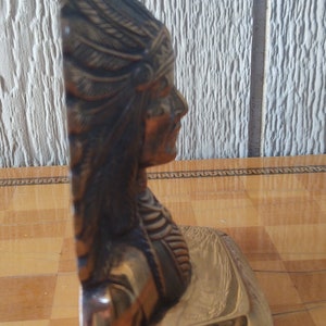 Puede incluir: Escultura en tono bronce de un jefe nativo americano de perfil. La escultura presenta un tocado de plumas y rasgos faciales detallados. La base es de color dorado pulido. Es probable que sea un objeto decorativo o de colecci&oacute;n.