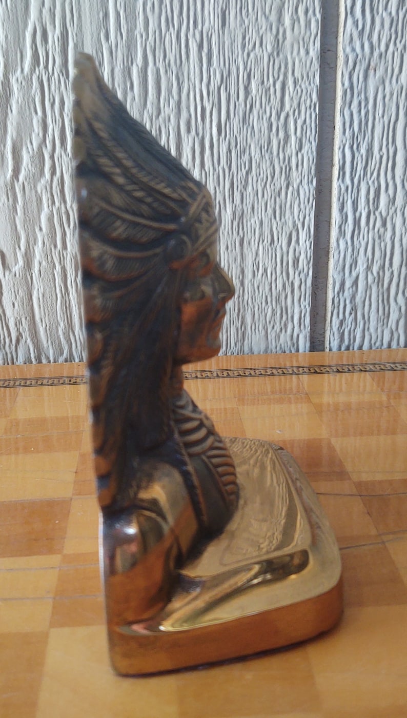 Puede incluir: Escultura en tono bronce de un jefe nativo americano de perfil. La escultura presenta un tocado de plumas y rasgos faciales detallados. La base es de color dorado pulido. Es probable que sea un objeto decorativo o de colecci&oacute;n.