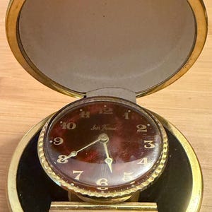 May include: A round gold-tone travel alarm clock with a hinged lid. The clock face features a deep red marbled background with gold-colored Arabic numerals and hands. The text Seth Thomas is printed in white script in the center of the clock face.
