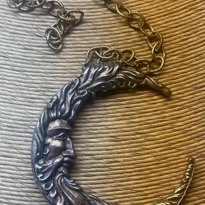 May include: A silver-toned crescent moon pendant with a detailed face and flowing hair design. The pendant is attached to a gold-colored chain. The necklace is displayed on a textured, light brown surface.