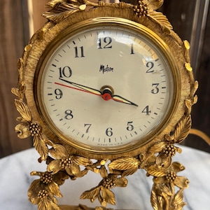 May include: A gold-toned metal table clock with a round face, black numerals, and the word Motion printed in the center. The clock features a decorative frame with sculpted leaves, flowers, and a bird perched on top. It has black hour and minute hands and a red second hand.