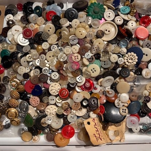May include: A large collection of assorted sewing buttons in various shapes, sizes, and colors, including plastic, metal, and wood materials. The buttons are loose and attached to cardboard backing cards, with one card featuring the text Lans.