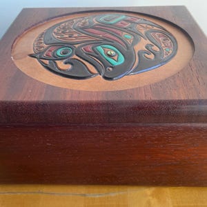 May include: A rectangular wooden box with a dark brown finish. The lid features a circular inset panel made of lighter brown material, decorated with a black, red, and turquoise graphic design depicting a stylized animal figure.
