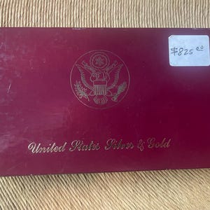 May include: A rectangular burgundy box with gold lettering and a gold emblem. The text "United States Silver & Gold" is visible. A white sticker with the price $825.00 is on the upper right corner.