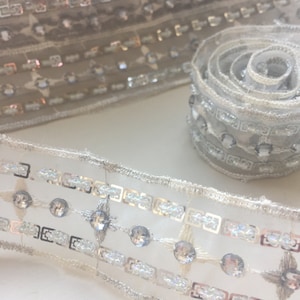May include: Silver and white beaded trim with a geometric pattern. The trim is made of a sheer fabric with small silver squares and round beads.