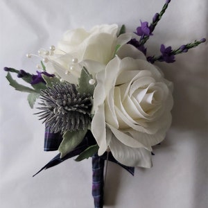 Puede incluir: Un boutonniere de rosa blanca con un cardo y flores moradas, adornado con cuentas de perlas y una cinta a cuadros.