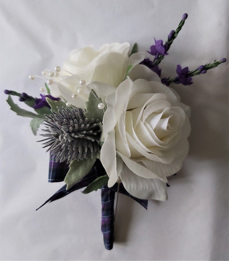 Puede incluir: Un boutonniere de rosa blanca con un cardo y flores moradas, adornado con cuentas de perlas y una cinta a cuadros.