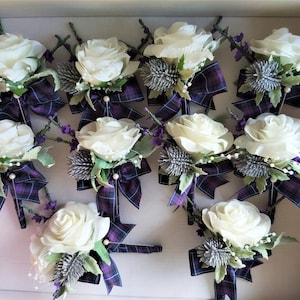 Puede incluir: Doce boutonni&egrave;res de rosas blancas con cinta a cuadros morado y verde y detalles de cardo plateado.
