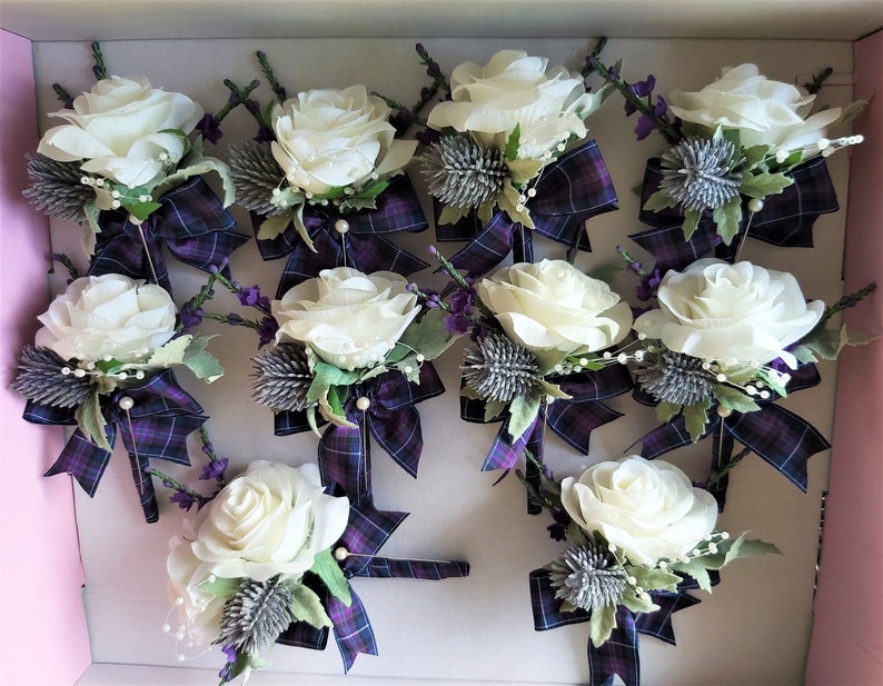 Puede incluir: Doce boutonni&egrave;res de rosas blancas con cinta a cuadros morado y verde y detalles de cardo plateado.