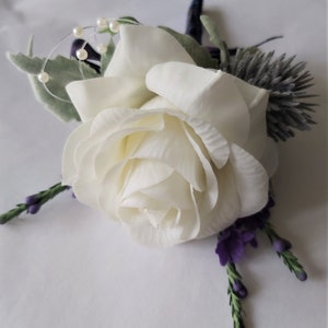 Puede incluir: Un boutonniere de rosa blanca con flores moradas y detalles de perlas.