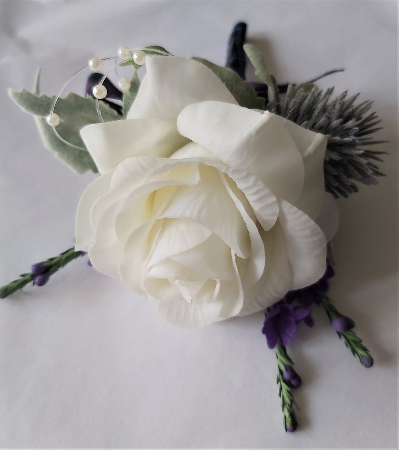 Puede incluir: Un boutonniere de rosa blanca con flores moradas y detalles de perlas.