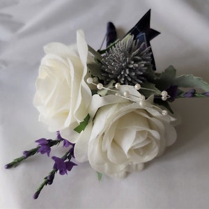 Puede incluir: Un boutonniere de rosa blanca con flores moradas, un cardo plateado y detalles de perlas.