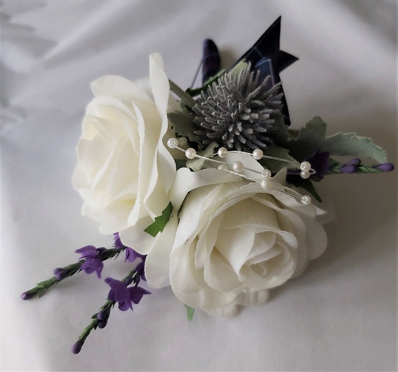 Puede incluir: Un boutonniere de rosa blanca con flores moradas, un cardo plateado y detalles de perlas.