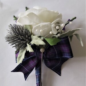 Puede incluir: Un boutonniere de rosa blanca con un lazo de tart&aacute;n morado y azul, un alfiler plateado y detalles de perlas.