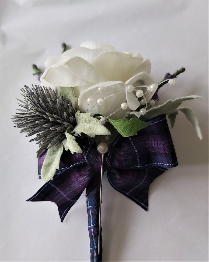 Puede incluir: Un boutonniere de rosa blanca con un lazo de tart&aacute;n morado y azul, un alfiler plateado y detalles de perlas.