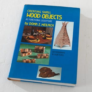 May include: A blue book titled "Creating Small Wood Objects" by Dona Z. Meilach. The cover features images of wooden sculptures, including a teepee-shaped structure and various decorative objects. The book is on a white surface.