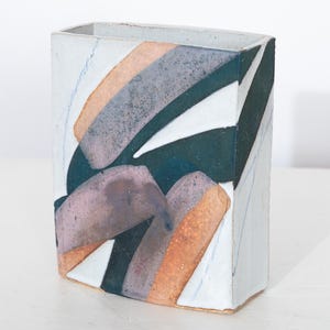 May include: Rectangular ceramic vase with abstract design. The vase features a white base with overlapping shapes in shades of gray, orange, and teal. The vase has a rectangular shape with a slightly angled base and a rectangular opening at the top.