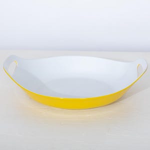 May include: A bright yellow serving dish with a white interior. The dish has a curved, oval shape with two white handles on either side. The dish is sitting on a white surface.
