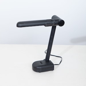 May include: A black desk lamp with a flexible arm and a rectangular base. The lamp is turned on and the light is shining.