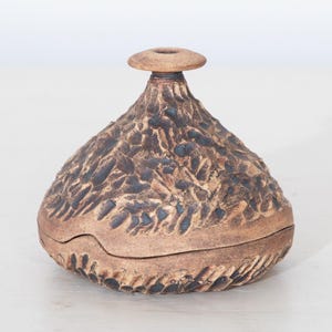 May include: A small, round, brown ceramic lidded vessel with a textured, carved surface featuring dark, recessed markings. The container has a wide, bulbous body that tapers to a narrow neck and a flat, circular rim at the top opening.