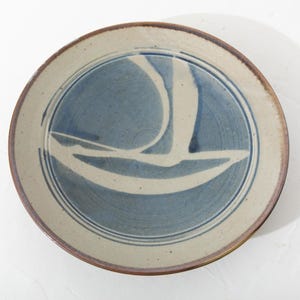 May include: Round ceramic plate with a blue and white sailboat design. The plate has a beige rim and a brown edge. The sailboat design is in the center of the plate, with a blue background and white lines.