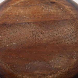 May include: A close-up of the bottom of a wooden bowl. The bowl is a warm brown color with visible wood grain and some light scratches. The text "DANSK DESIGN DENMARK" is stamped into the wood.