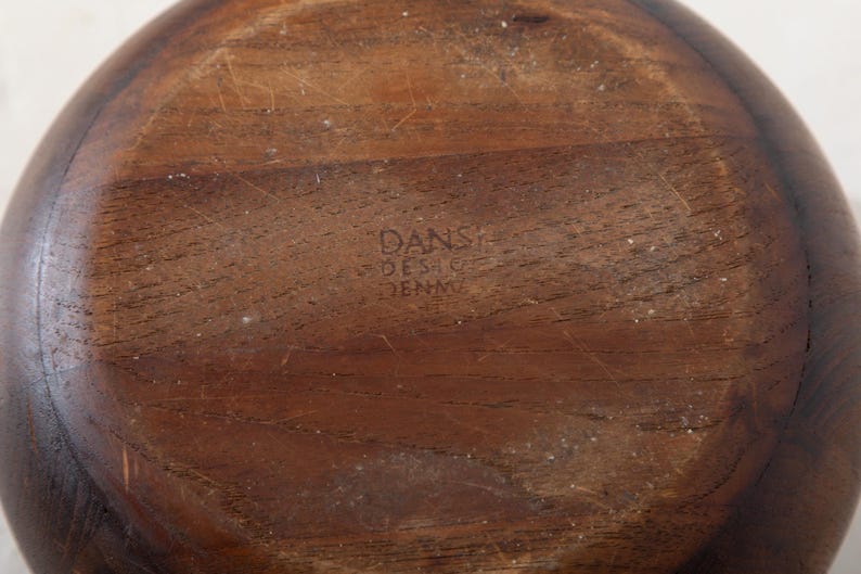 May include: A close-up of the bottom of a wooden bowl. The bowl is a warm brown color with visible wood grain and some light scratches. The text "DANSK DESIGN DENMARK" is stamped into the wood.