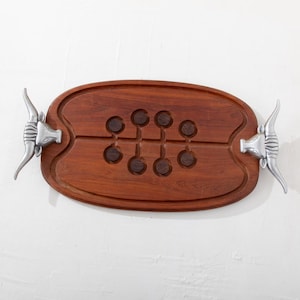 May include: A wooden serving board with a dark brown finish and a football shape. The board has six circular indentations and is adorned with silver-colored bull head handles on each end. A decorative piece for serving.