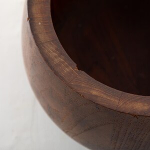 May include: Close-up of a wooden bowl with a dark brown finish. The bowl's rim is visible, showing the natural wood grain and a slight imperfection. The interior is dark, suggesting a deep cavity. This handcrafted bowl is suitable for kitchen use.